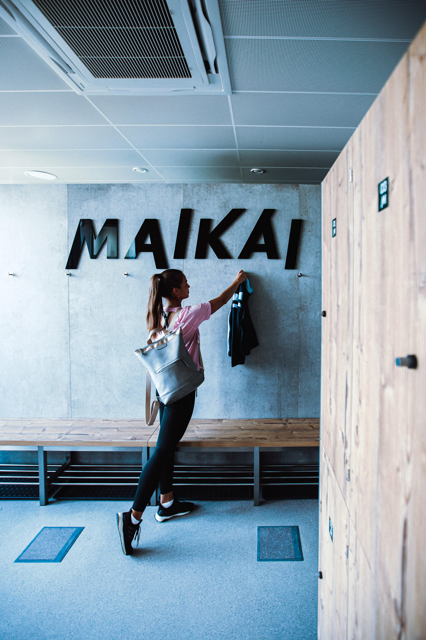 Frau mit Rucksack die ihre Jacke an Hacken in einer Umkleide hängt. An der Wand der Schriftzug "MAIKAI"