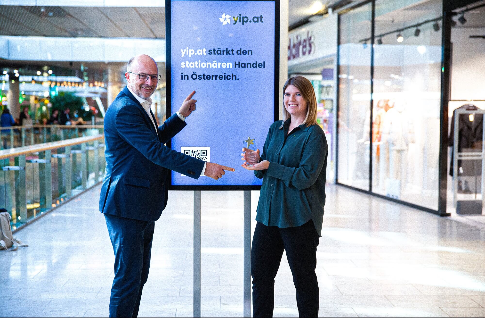 Frau mit grüner Bluse und European Solal Award in der Hand und Mann im Anzug der auf eine Werbeanzeige zeigt auf der steht "yip.at stärkt den stationären Handel in Österreich"